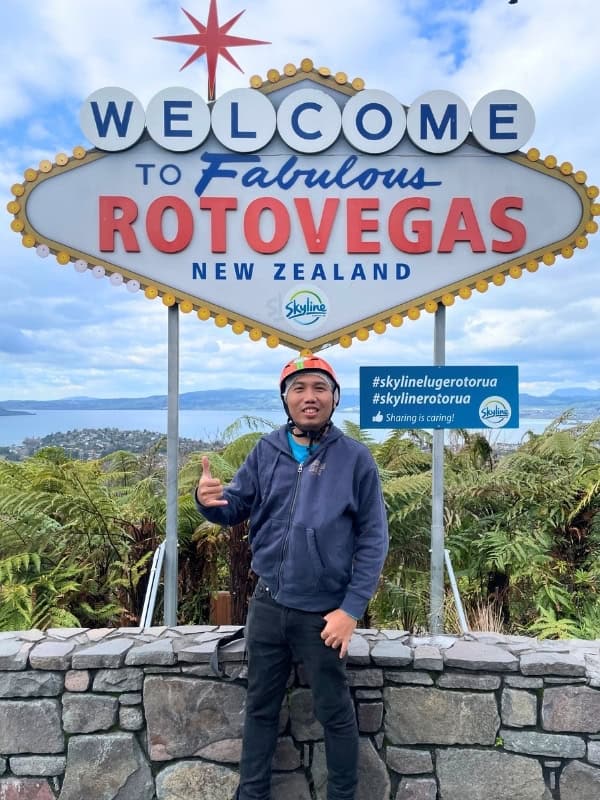 Traveler giving a thumbs-up beneath the Welcome to Fabulous Rotovegas sign overlooking Rotorua, New Zealand.