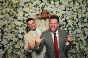 Groom and his friends having fun at a photo booth HD with floral backdrop, holding drinks, and making silly.