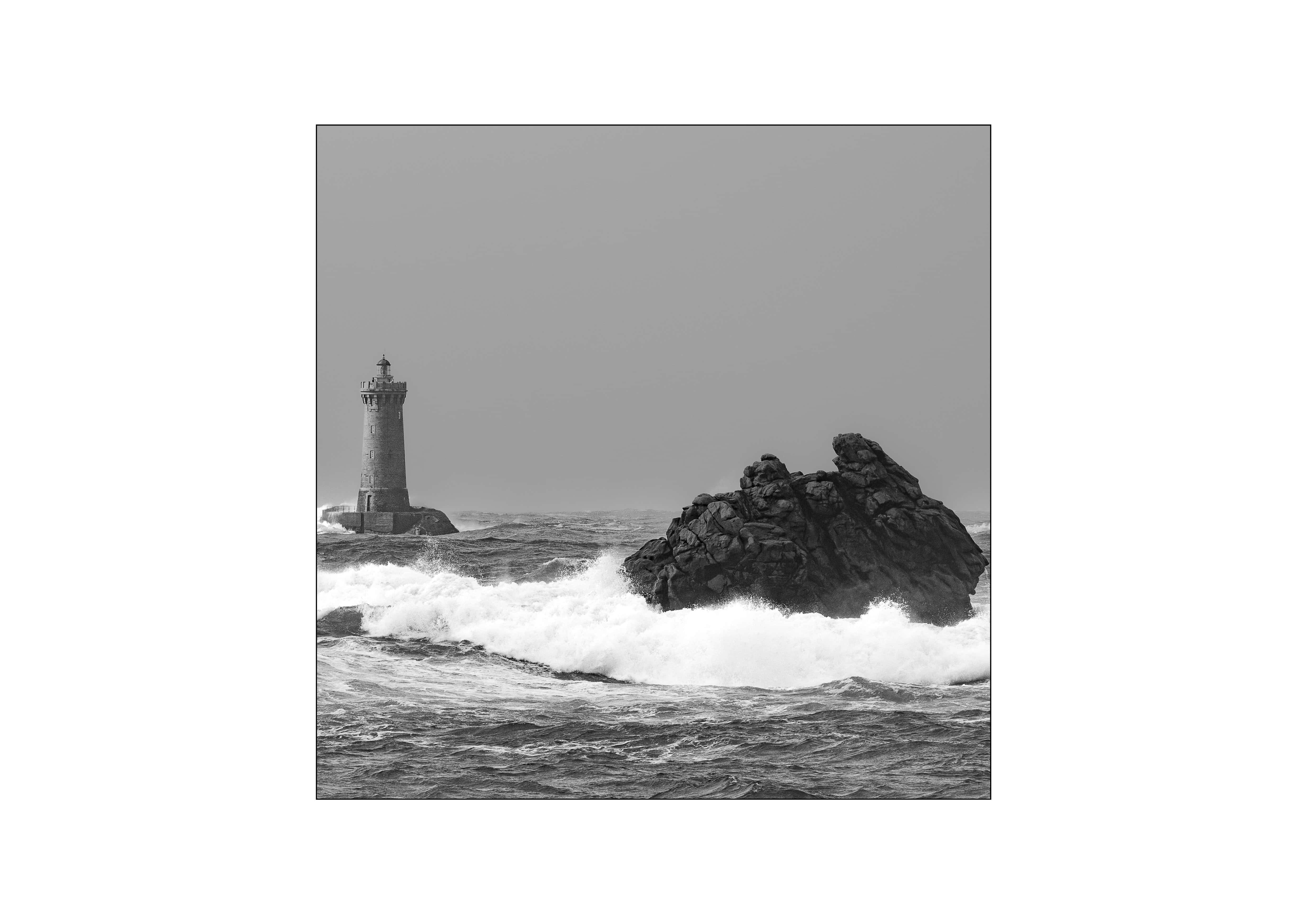 Black and white square photograph of the Phare du Four lighthouse off Le Conquet, Finistère, Brittany, with a large Atlantic storm wave breaking against rocks in a raging sea.