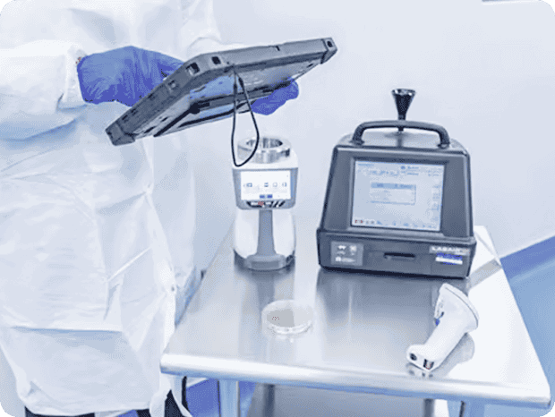 A person in cleanroom garments holding a tablet near digital monitoring equipment on a stainless‑steel table, including a particle counter and handheld scanner.