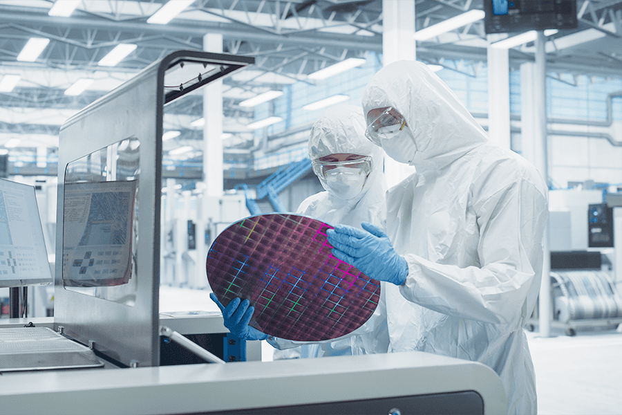 Technicians in cleanroom gear inspecting a semiconductor wafer beside computer equipment on the production floor.