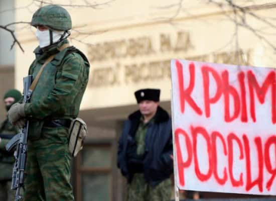 Військовий у захисному спорядженні з автоматом на фоні протесту з плакатом про Крим.