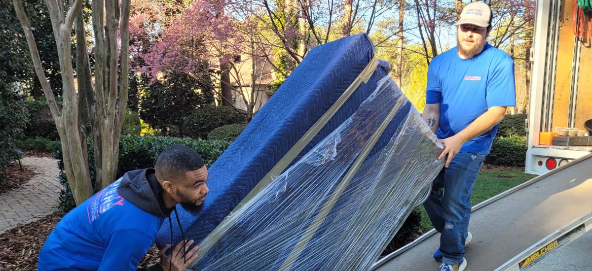 Two Port City Movers team members moving a sofa wrapped in protective covering up a ramp.