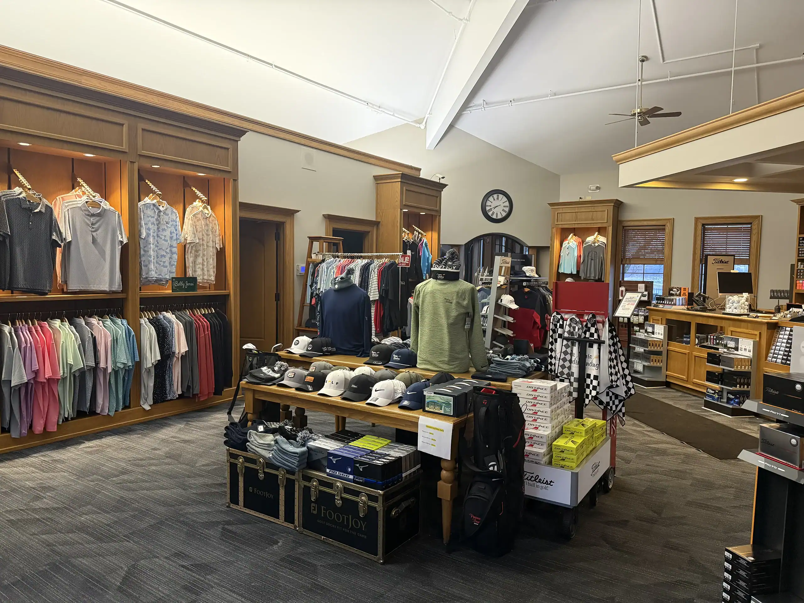 Interior view of a well-stocked golf pro shop, featuring wooden displays with golf apparel, hats, golf balls, and accessories. A central table holds hats and a mannequin in a green golf shirt.