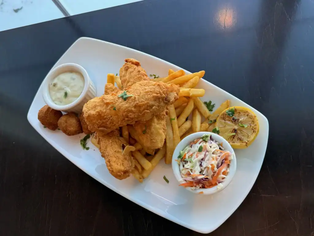 A white rectangular plate filled with golden-brown fried fish, French fries, a side of creamy tartar sauce, three hush puppies, a bowl of colorful coleslaw, and a grilled lemon half, all on a dark table.