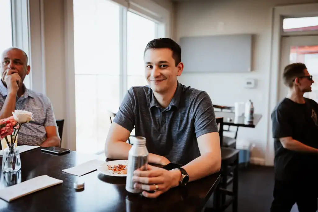 A young man smiles at the camera while holding a metal bottle at a table, with an older man and another person in the background.