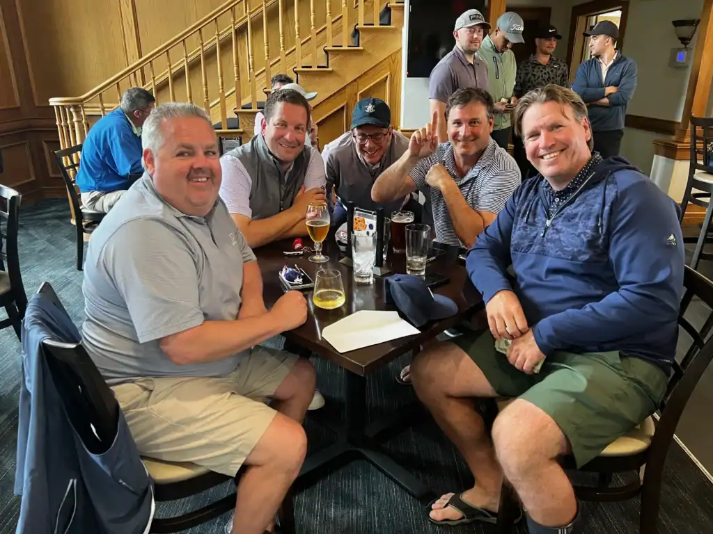 A group of smiling men, dressed in golf attire, sit around a table with drinks in a clubhouse, celebrating after a game. One man holds cash and makes a 'number one' gesture.