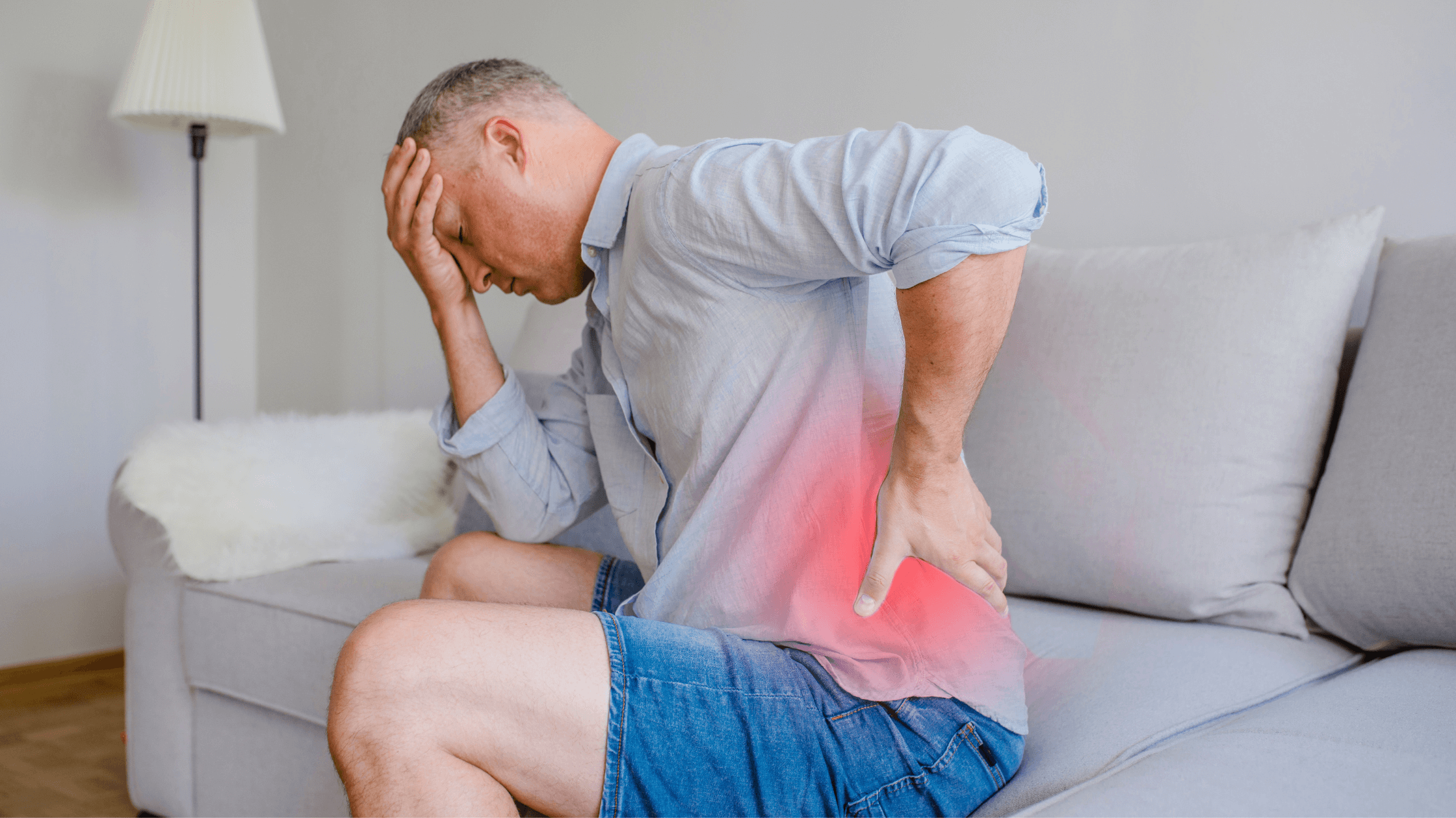 A man sits on a sofa, holding his lower back with a red glow highlighting the painful sacroiliac joint region, illustrating the discomfort of SI joint pain that injections can help relieve.