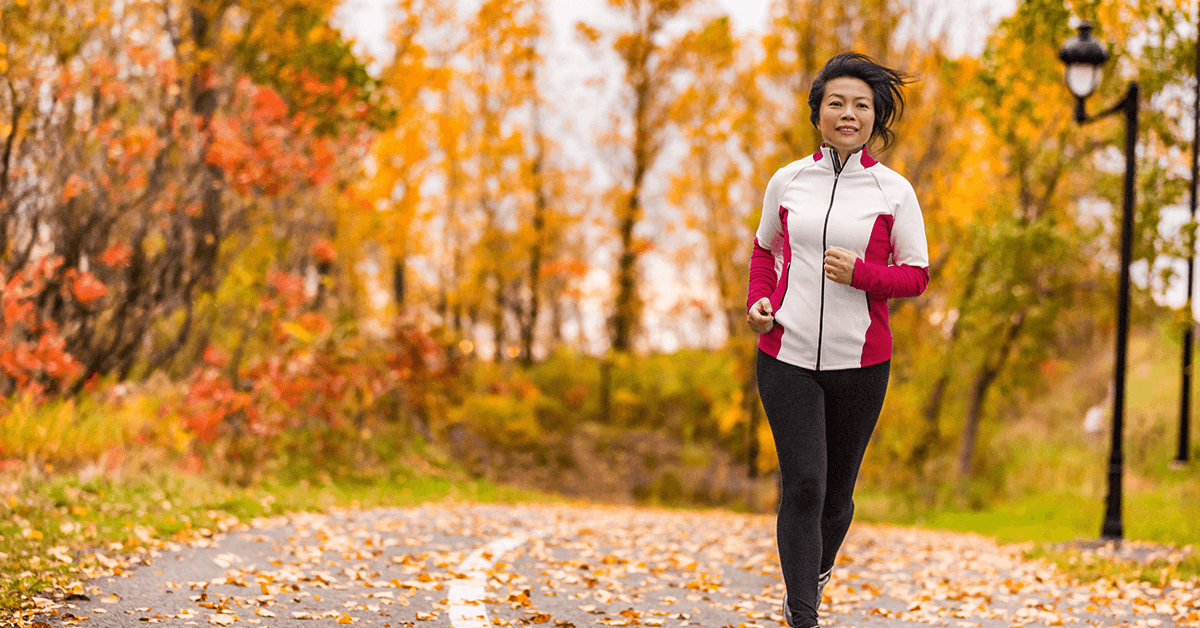 A woman is jogging outdoors on a path covered in autumn leaves, wearing a white and red athletic jacket, symbolizing improved mobility, fitness, and an active lifestyle achieved after successful pain management treatment.