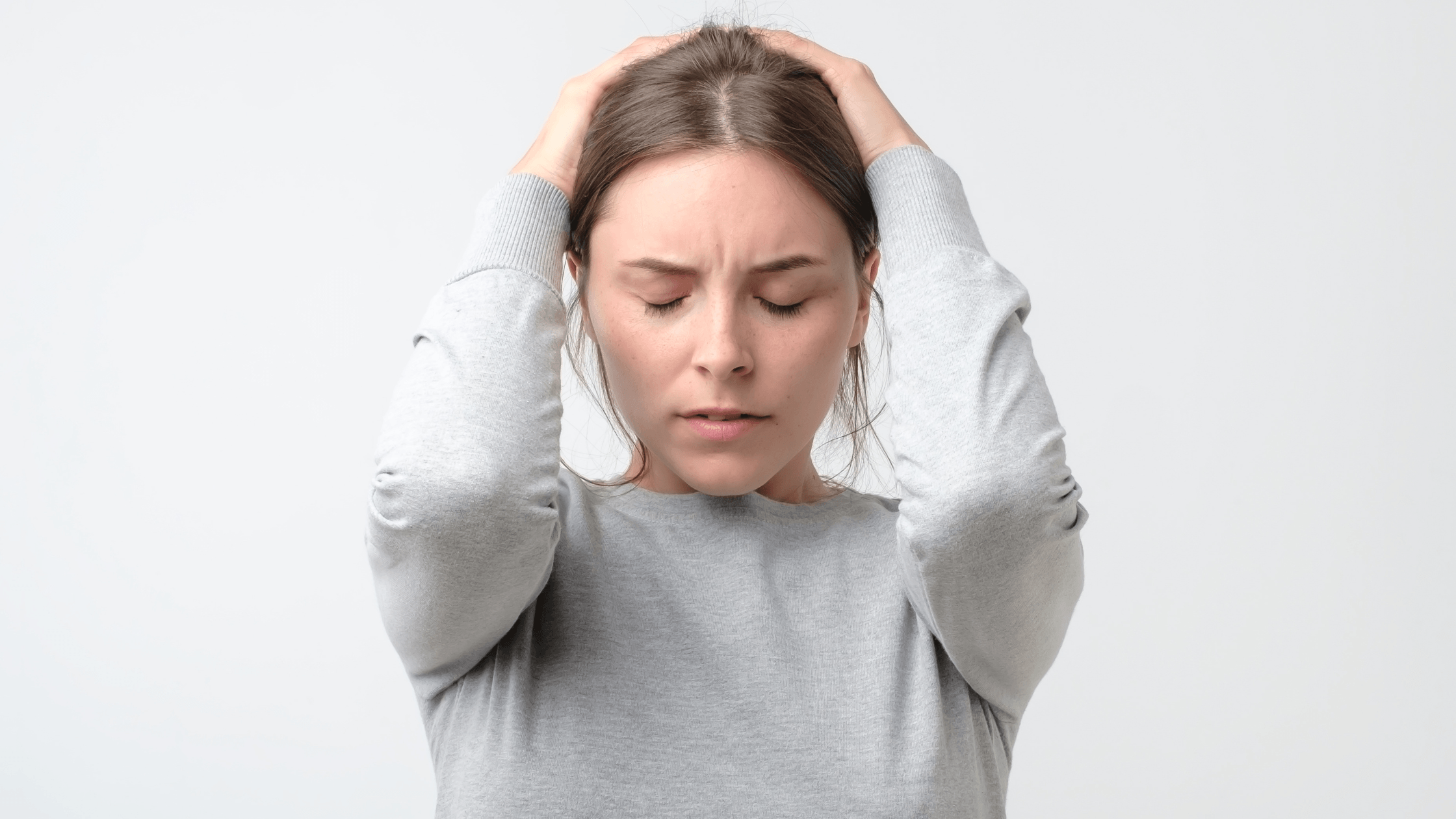 A woman in a grey sweatshirt holding her head with both hands, eyes closed in an expression of intense pain or a severe headache.
