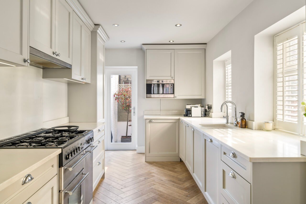 Modern white kitchen with natural light, wooden flooring, and stainless steel appliances.