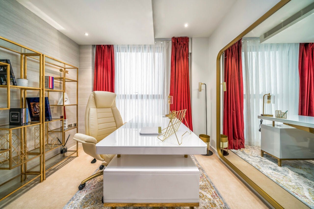 Modern office with white desk, beige chair, gold shelving, and red curtains.