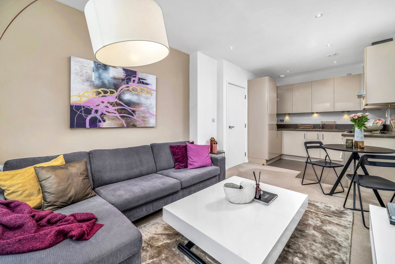 Modern open-plan living room with grey sofa, colourful cushions, and kitchen area.