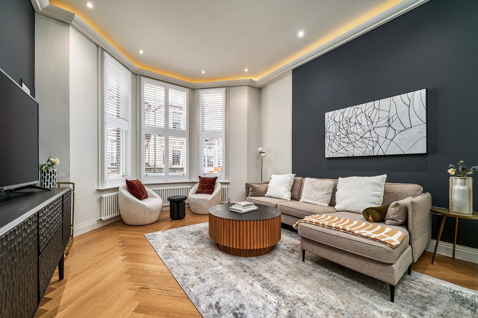 A modern living room with large bay windows, grey accent wall, and stylish furniture.