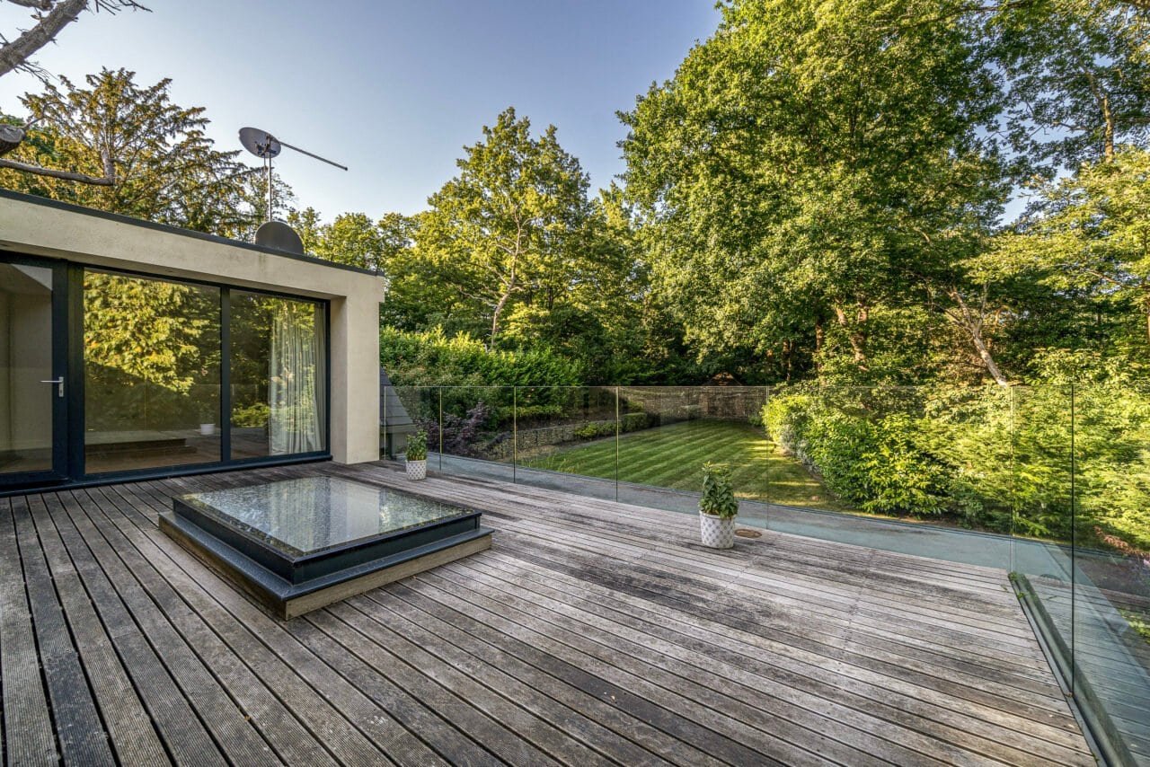 Modern wooden deck with glass railing and lush green garden in the background.