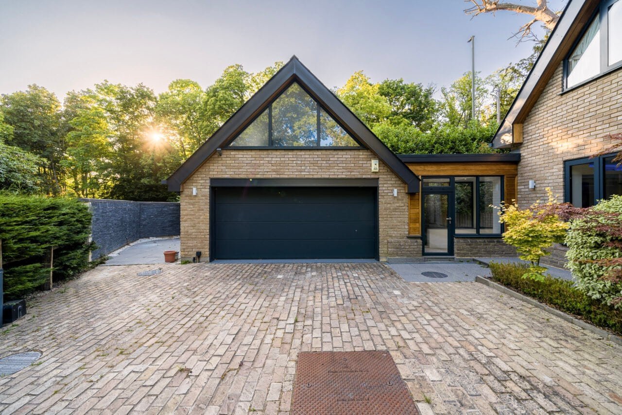 A modern detached house with a brick driveway and garage, surrounded by greenery.