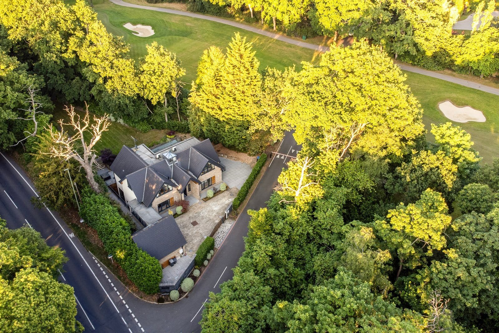 Aerial view of a modern house surrounded by lush green trees and a golf course in the background.