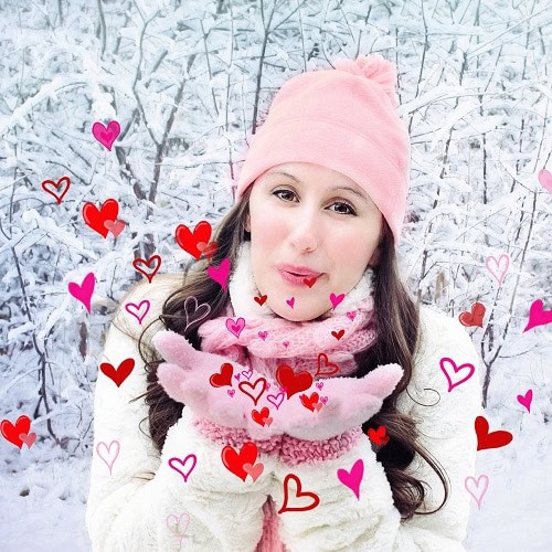 A woman in winter clothing blowing snowflakes with heart shaped overlays.