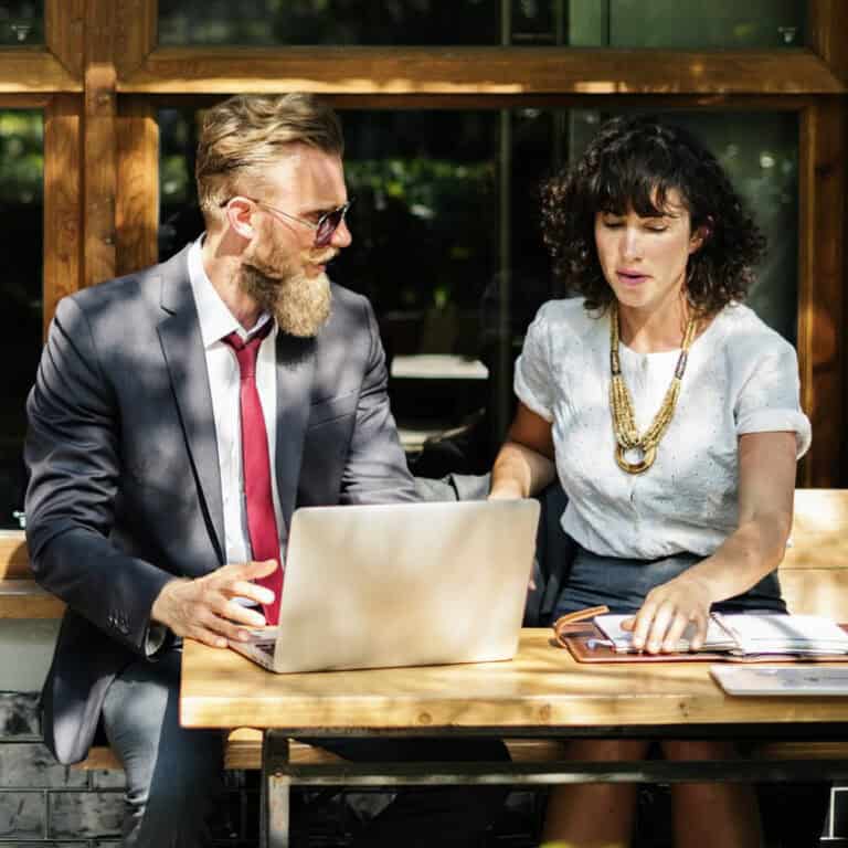 Two professionals collaborating outdoors at a wooden table with a laptop.