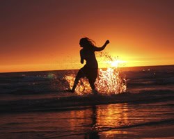 Woman running along the beach at sunset with waves splashing around her.