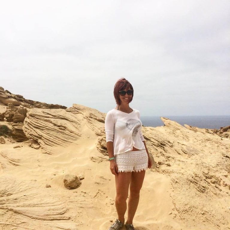 Beach landscape with a woman in a white dress and sunglasses standing on sandy rocks.
