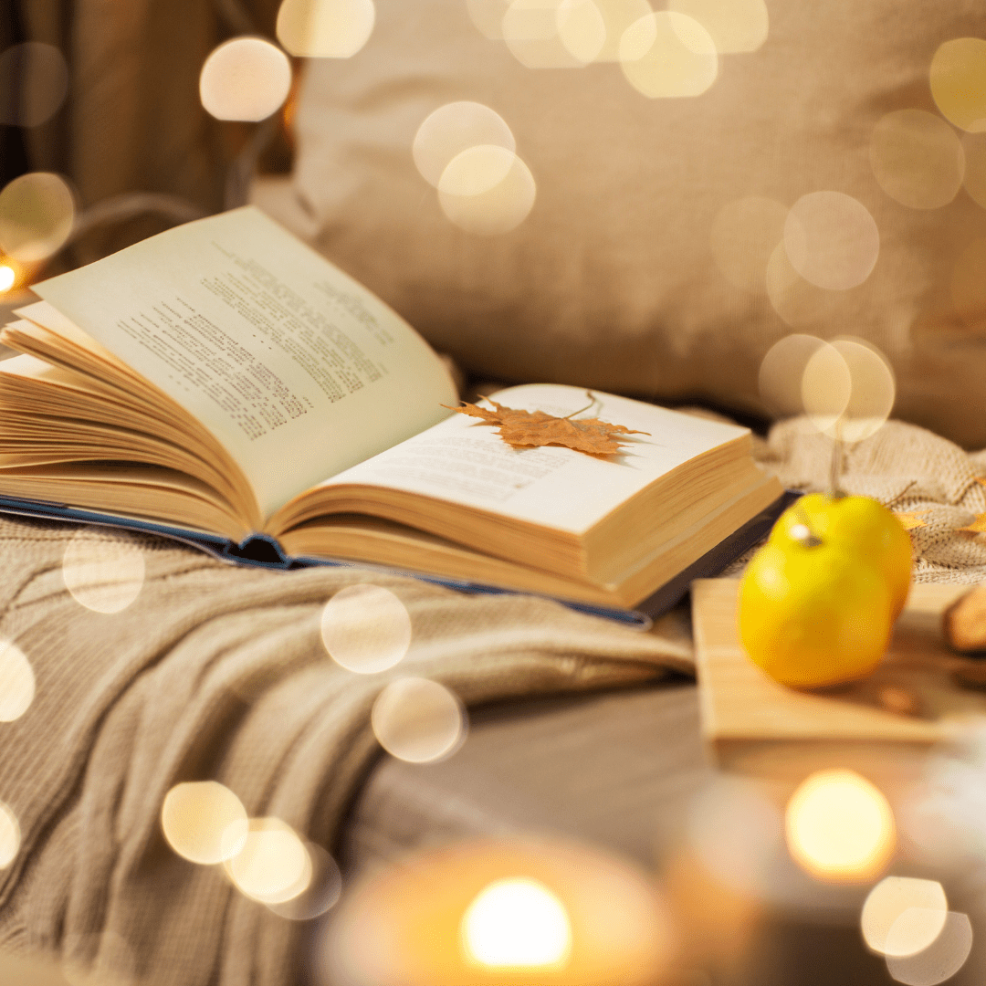 Open book on cozy blanket with warm lights, autumn leaf, and pear.