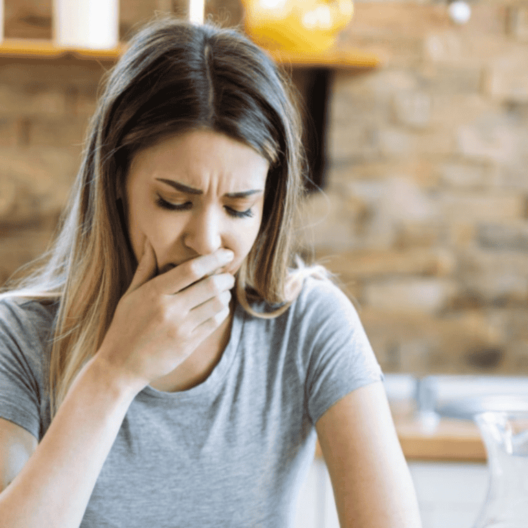 Woman experiencing nausea in kitchen, holding her hand over her mouth.