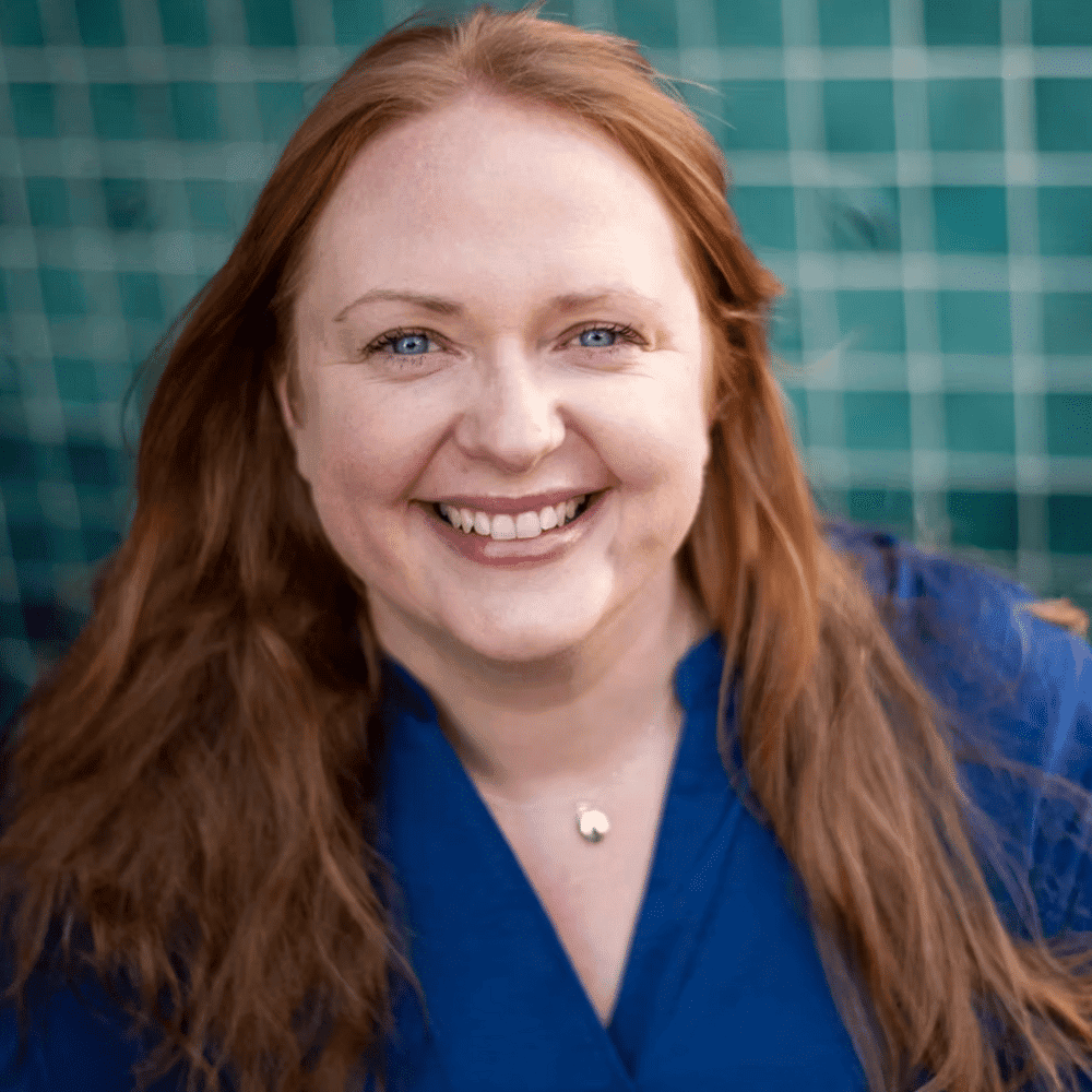 Healthy smiling woman with red hair in blue scrubs against a green tiled background.