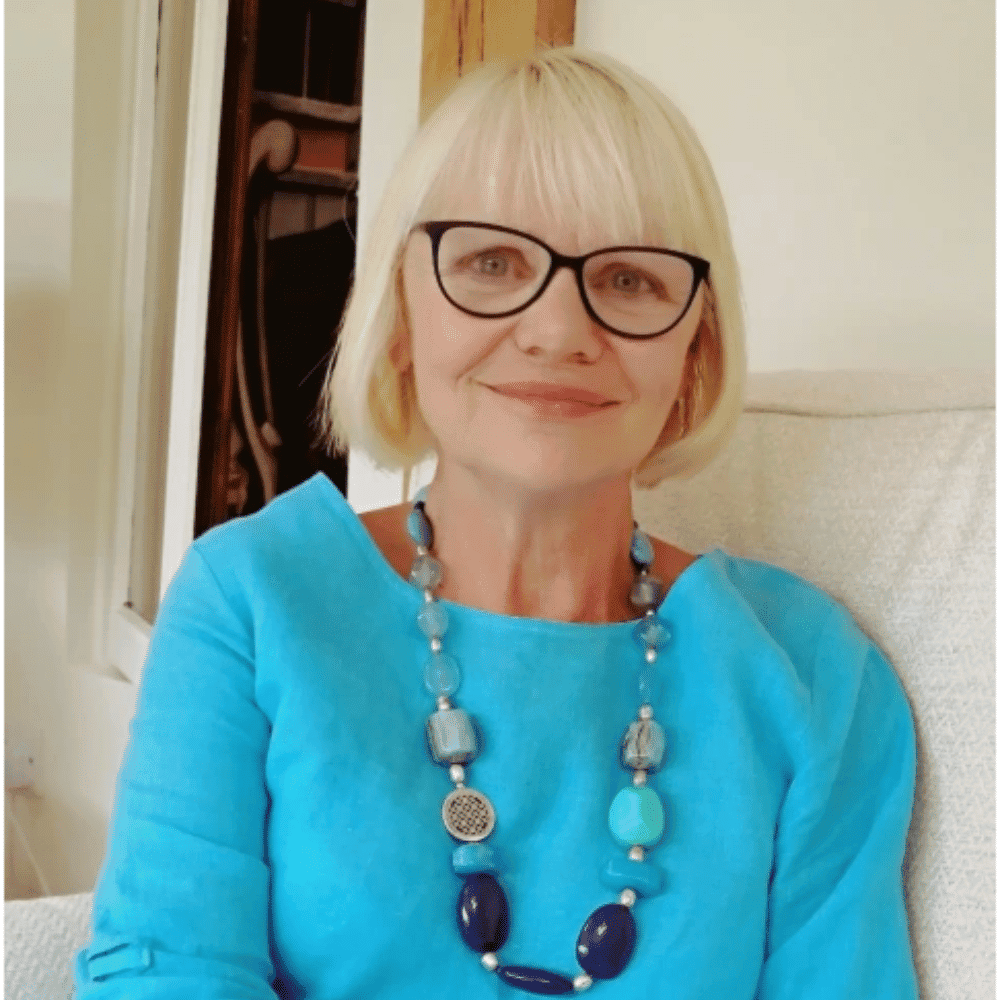 Elegant woman with blonde hair, glasses, and a vibrant blue top, smiling warmly indoors.