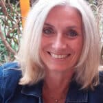 Smiling woman with blonde hair and blue eyes, wearing a denim jacket, in a natural outdoor setting.