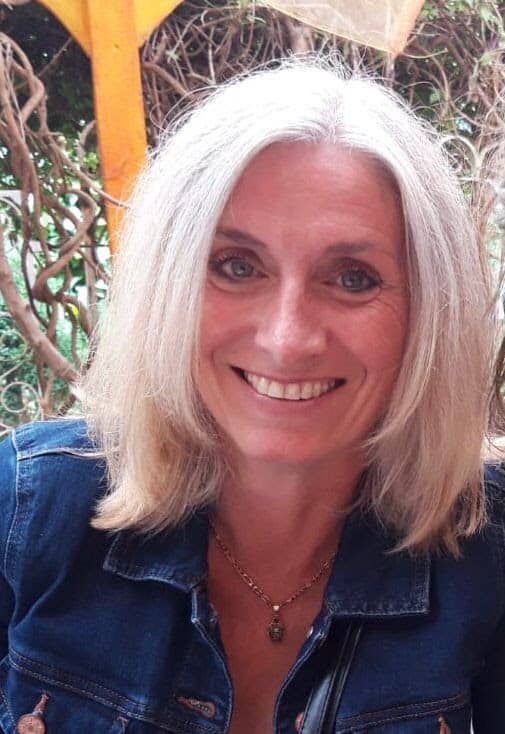 Smiling woman with blonde hair and blue eyes, wearing a denim jacket, in a natural outdoor setting.