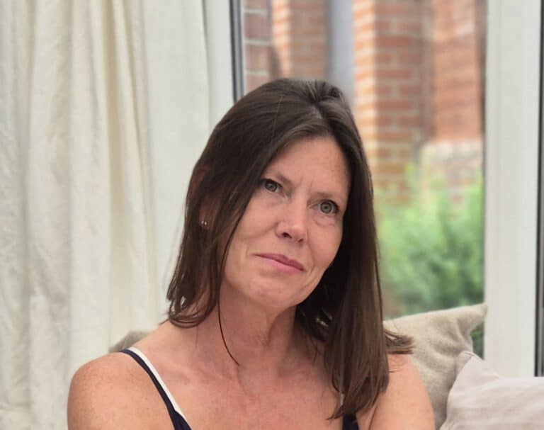 Woman with brown hair sitting indoors near a window, thoughtful expression.