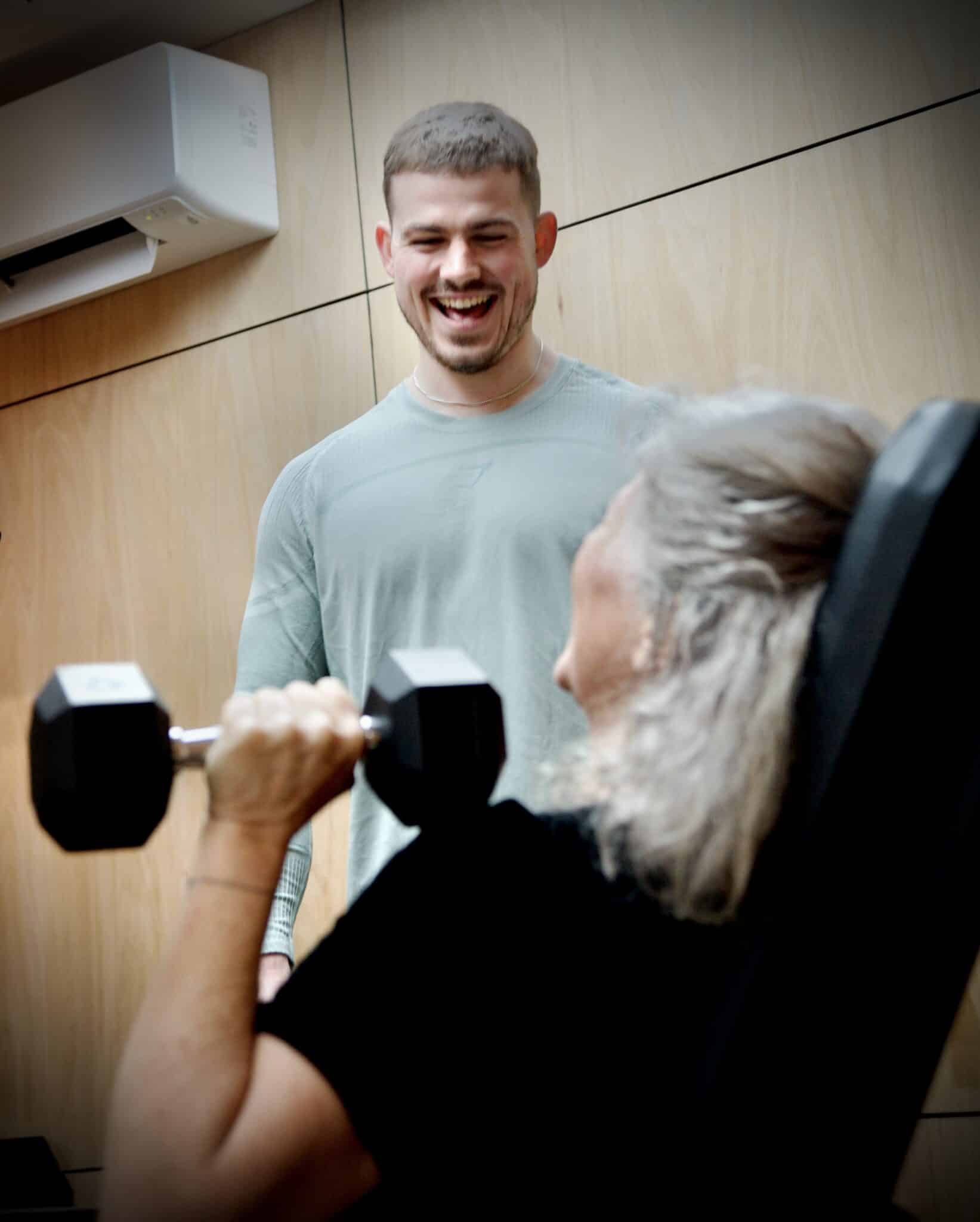 Elderly woman lifting dumbbell with a young trainer smiling nearby.