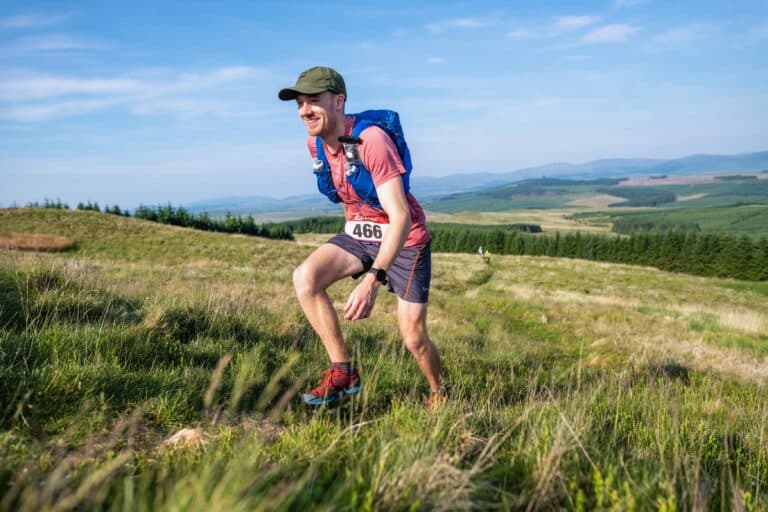 Hiker with backpack and race bib running through grassy landscape.