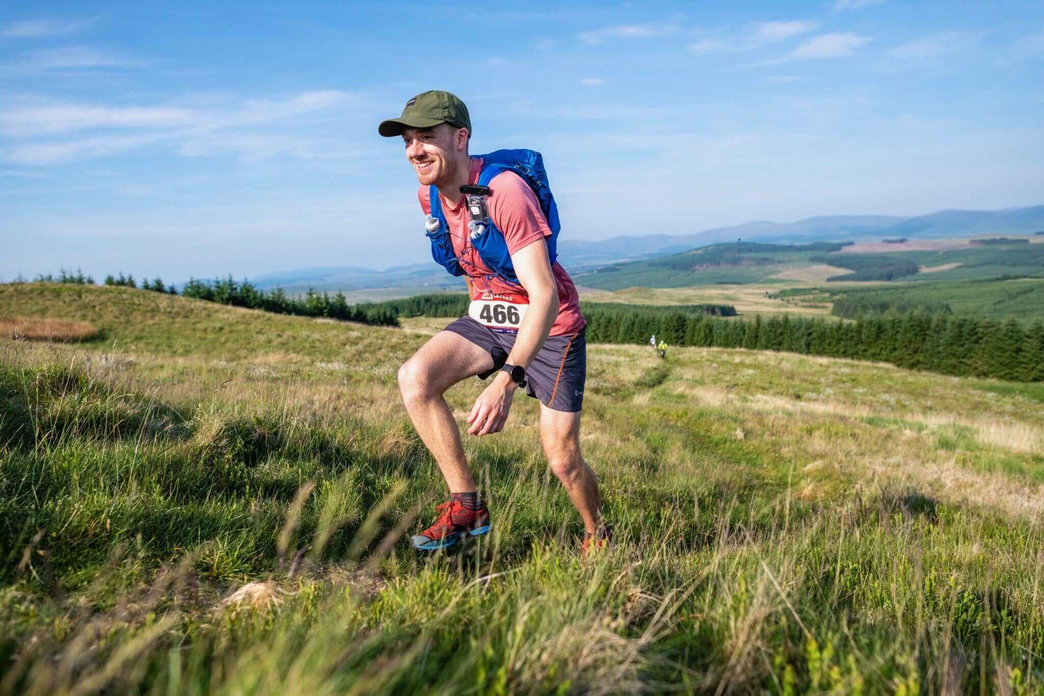 Hiker with backpack and race bib running through grassy landscape.