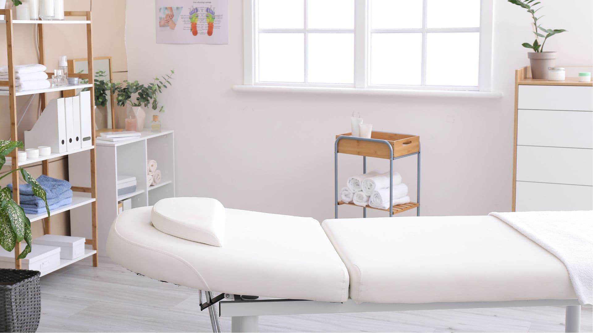 Bright, clean chiropractic treatment room with a massage table, shelves, plants, and natural light.