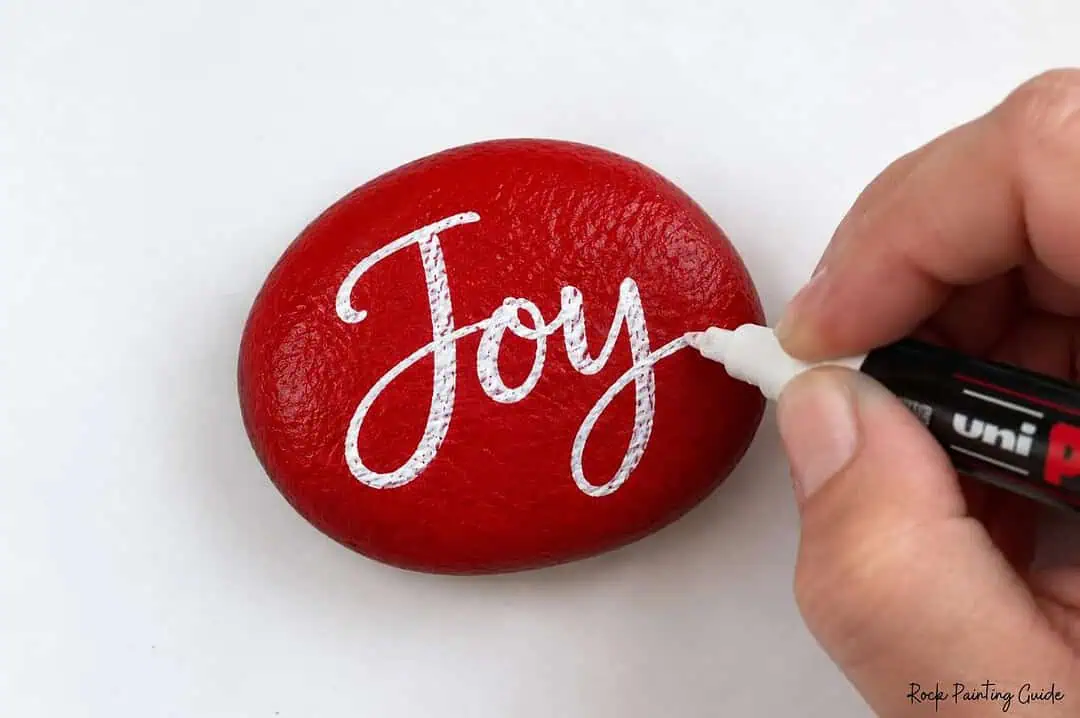 200+ Christmas Quotes and Sayings for Crafts (Free Printable) 8 Red rock with white handwritten word Joy being penned.