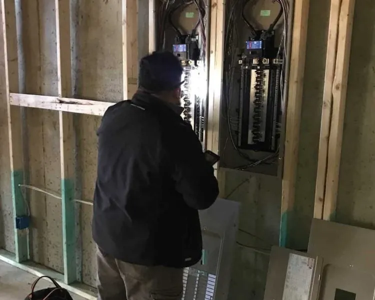 An NDI inspector examining an open electrical panel between the exposed studs of a house under construction