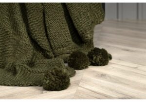 Mörkgrön stickad filt och små pompom-tyg på en ljus träsoffa.