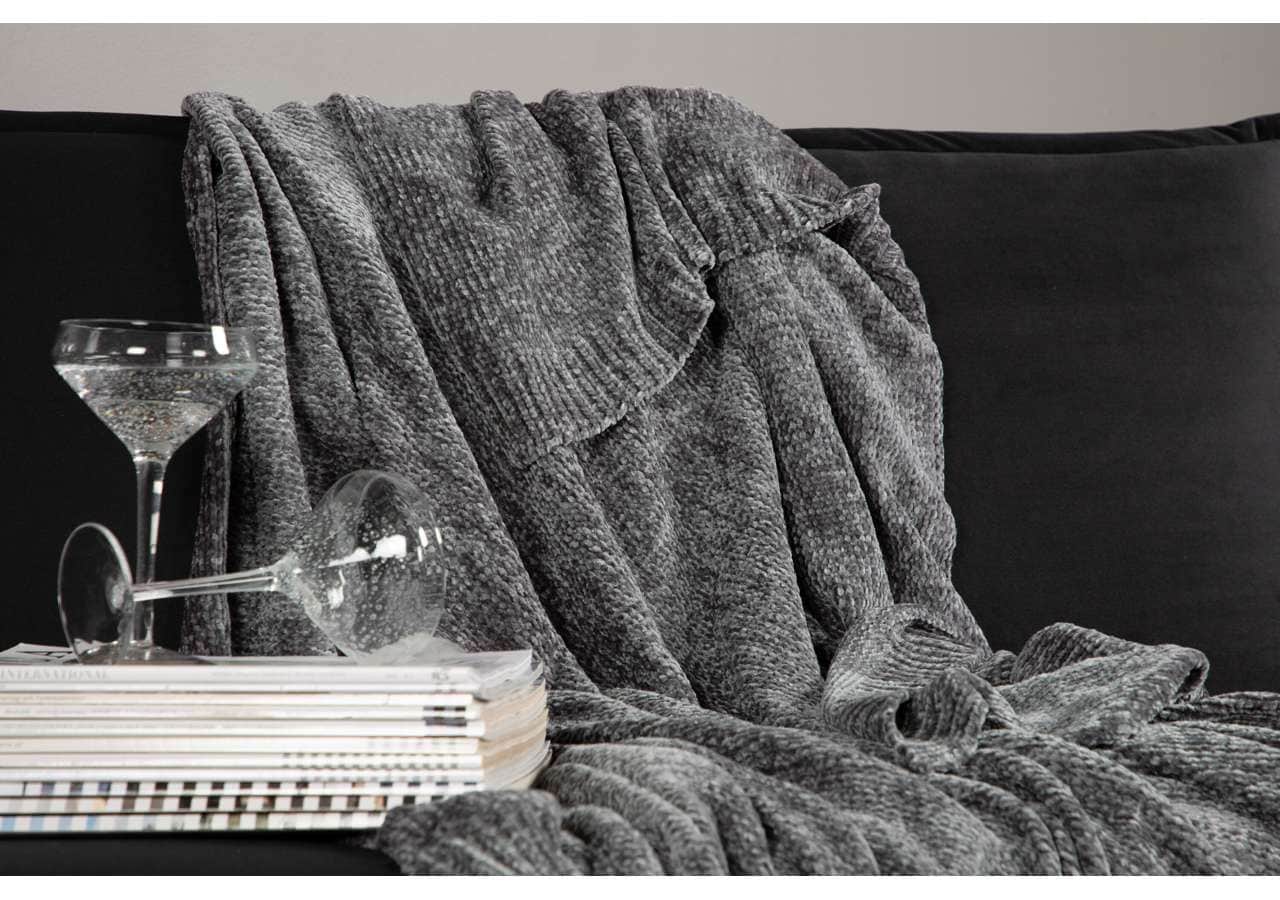 Mörkgrå stickad pläd på svart soffa med glasögon och böcker, perfekt för mys och avkoppling.