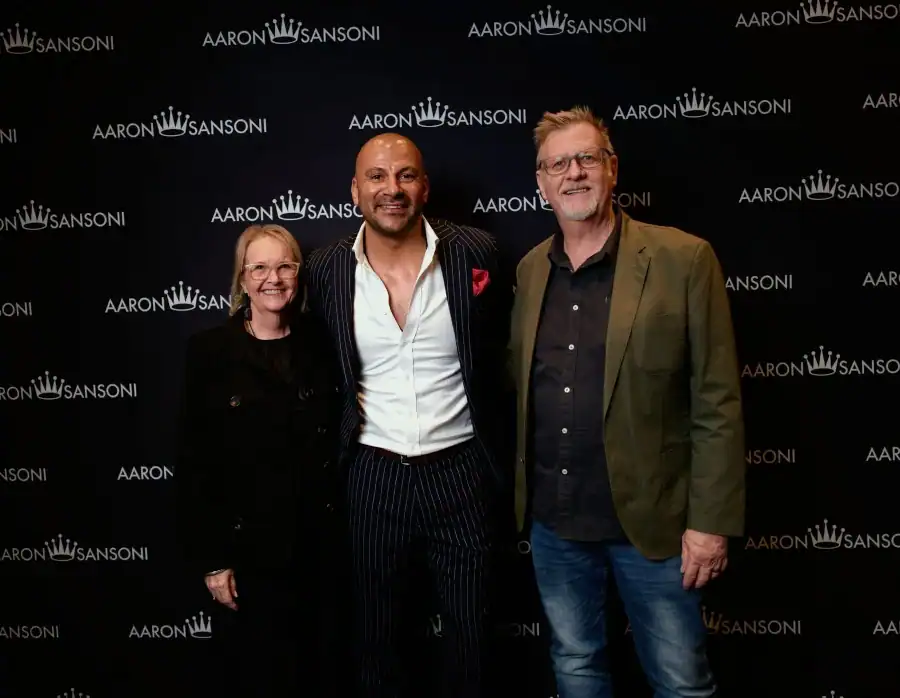 Three people stand smiling in front of a black backdrop with repeated AARON SANSONI logos. The person in the centre wears a striped suit; the two others wear glasses and casual outfits.
