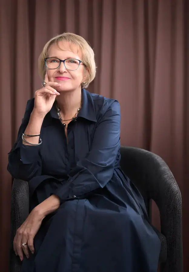 A woman with short blonde hair and glasses sits in a chair, wearing a dark blue dress, resting her chin on her hand and smiling thoughtfully. A brown curtain is in the background.