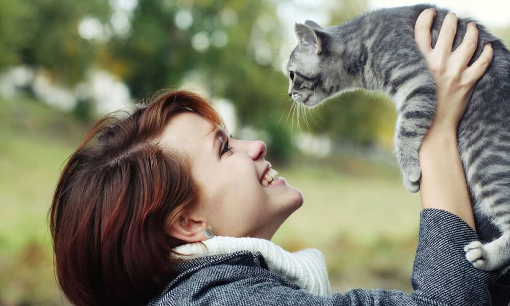 A joyful woman veterinarian with short brown hair holding a gray tabby kitten, smiling as they look at each other outdoors with blurred greenery in the background.