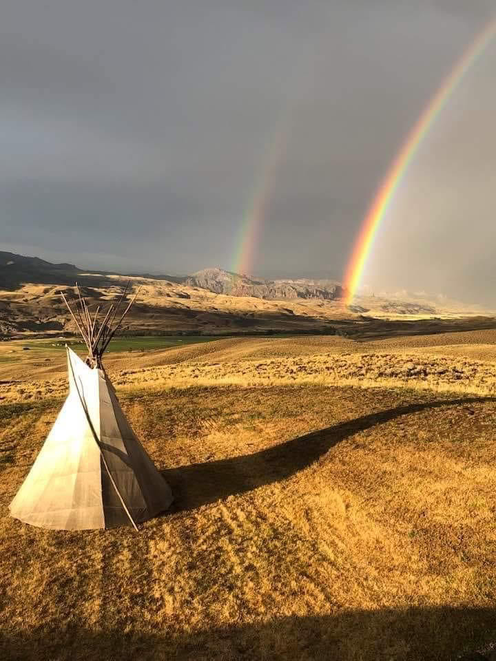 Rainbow over grassland with tipi tent and mountain backdrop, reflecting hope and spiritual renewal for Singing Feather Ministries.