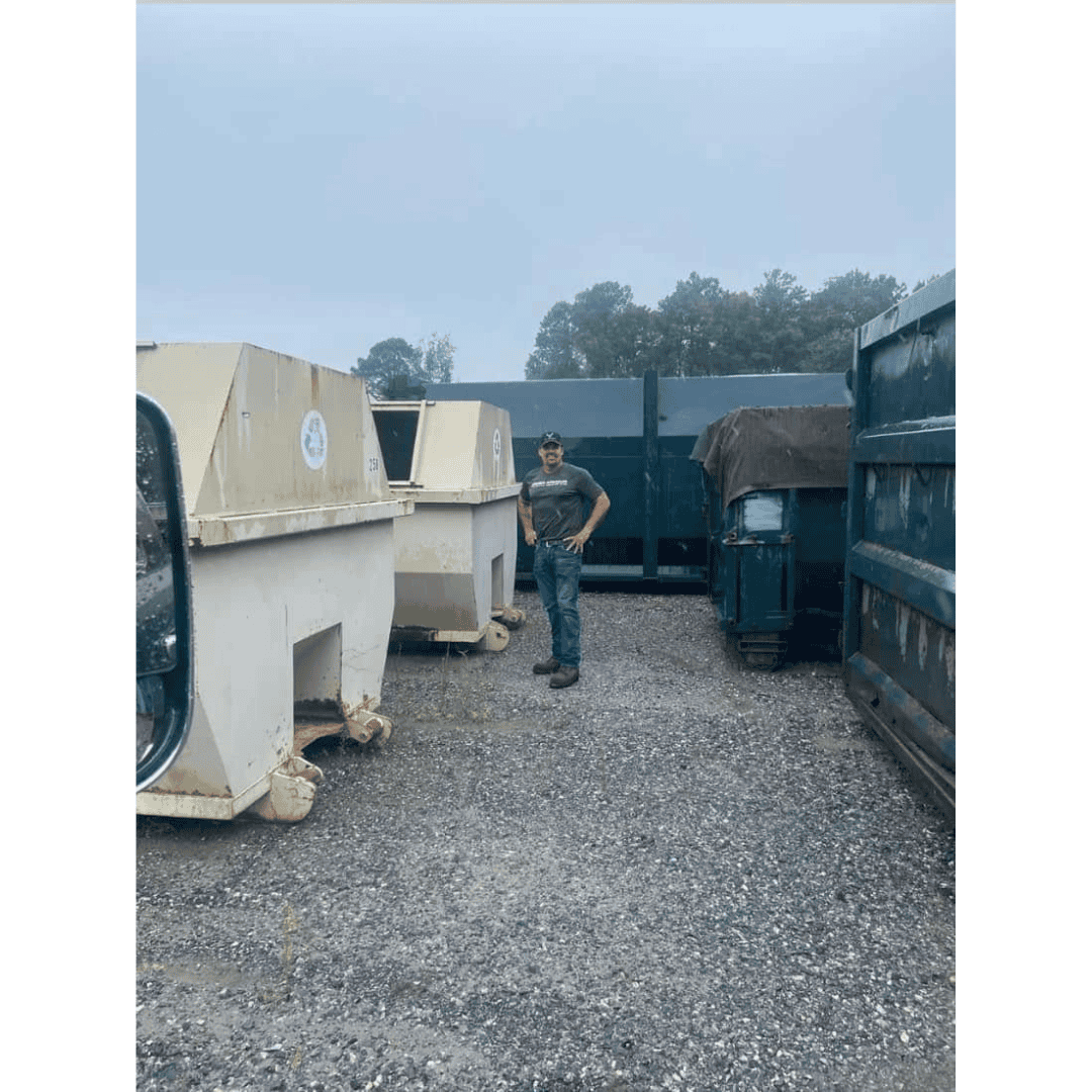 A person standing among waste containers and dumpsters in an outdoor storage yard.