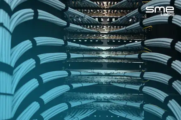 Pale grey-blue network cables gathered into strapped bundles curving into a tall patch panel rack lit warm gold down its centre, with SMEnode Labs cyan entering from the bottom-left corner and easing out diagonally toward the top right.