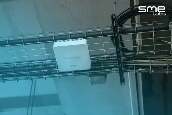 A single white network device mounted on a run of steel cable tray carrying bundled conduit across a concrete ceiling, with SMEnode Labs cyan entering from the bottom-left corner and easing out diagonally toward the top right.