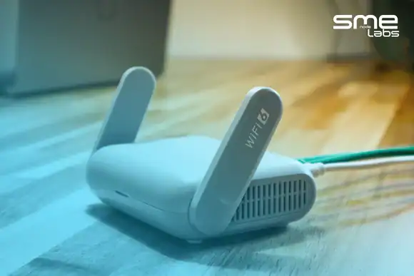 A white WiFi 6 home router on a sunlit wooden desk with a green cable plugged into its WAN port and a laptop behind it, with SMEnode Labs cyan entering from the bottom-left corner and easing out diagonally toward the top right.