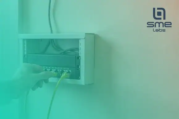A hand plugging a yellow network cable into a router inside a small wall-mounted network cabinet in an office, with SMEnode Labs teal fading in from the bottom-left corner.