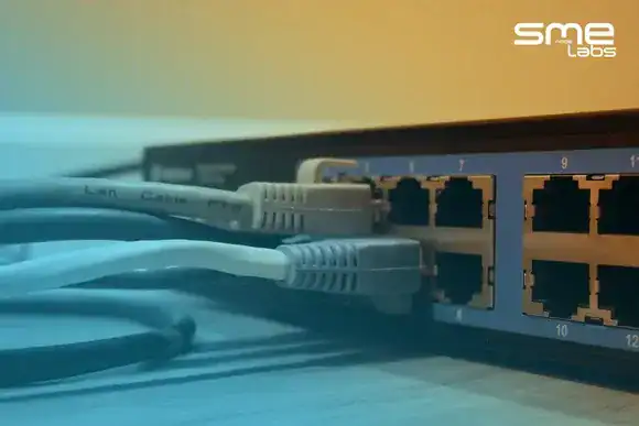 Patch cables plugged into the ports of a desk network switch, with SMEnode Labs cyan fading in from the bottom-left corner.